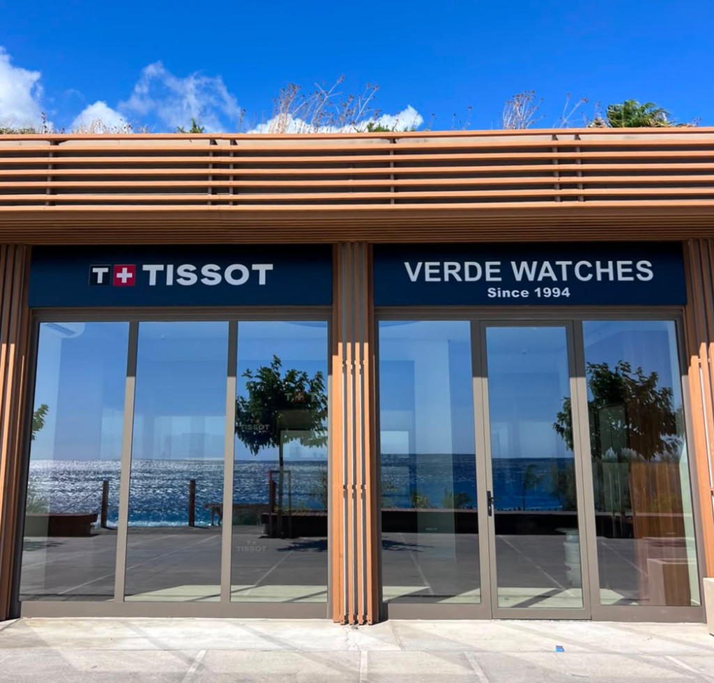 Seaside storefront with TISSOT and VERDE WATCHES signs, glass doors, and an ocean view.